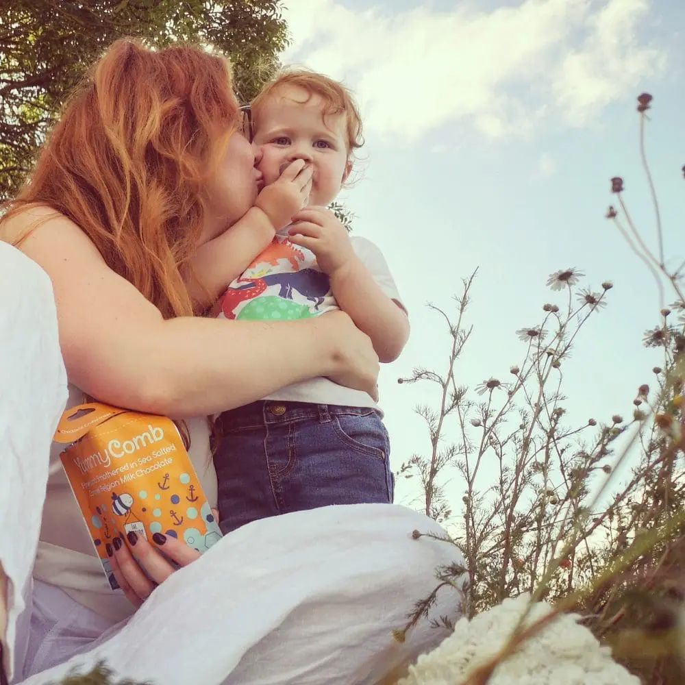 Mother and son sharing a pouch bag of salted caramel covered honeycomb in a field on a picnic rug